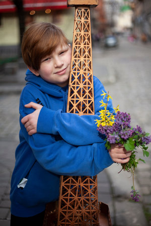 A boy with red hair and a blue sweatshirt with a bouquet of purple wildflowers hugs the Eiffel Tower
