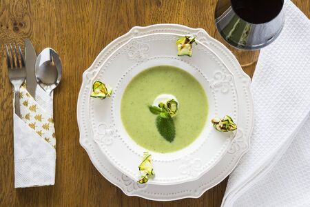 Broccoli cream soup in a porcelain plate with grilled zucchini slices, mint and cream and a glass of red wine on a wooden backgroundの写真素材