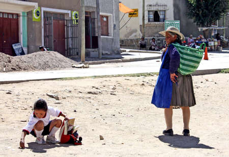 5 April, 2010 - La Quiaca, Bolivia- An old Bolivian woman waiting for the little schoolgirl who is picking up something that fell from her schoolbagのeditorial素材