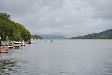 Calm morning at the lake with boats and mountains in the background during a cloudy dayの写真素材