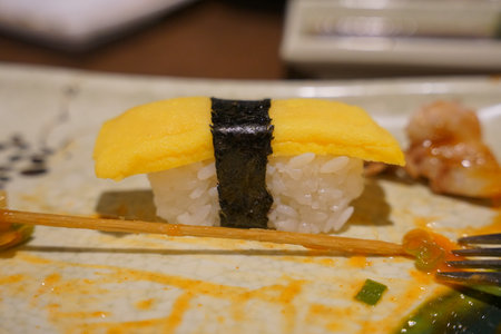 Unique sushi creation with yellow topping and seaweed wrap served on a decorative plate in a vibrant restaurant settingの写真素材