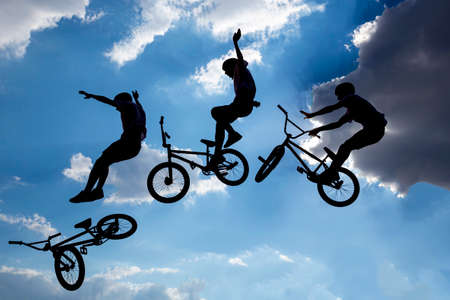 An extreme rider is making a free style jump from a ramp. The young boy with his bicycle is seen as a silhouette in front of the sun. Multiple exposure image. Clouds.の写真素材