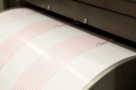 Seismograph records an earthquake on the sheet of measuring paper. Seismological device for measuring earthquakes. Seismograph machine needle drawing a red line on graph paper measuring activity.の写真素材