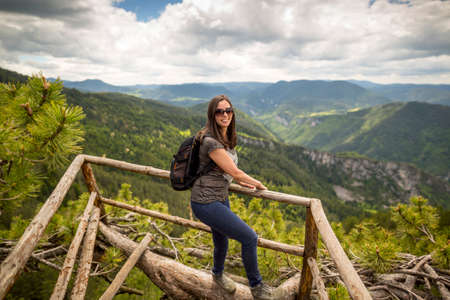 Young tourist woman with backpack enjoys the view from a wooden bridge high in the mountain designed for viewing the side from above. In pine trees. Vivid image of the mountain. Tourism destination.の写真素材