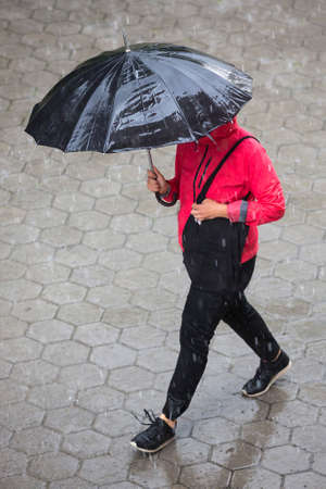 A woman runs in the rainy weather with her black umbrella. Raining like cats and dogs. Bad weather. Climate change.の写真素材
