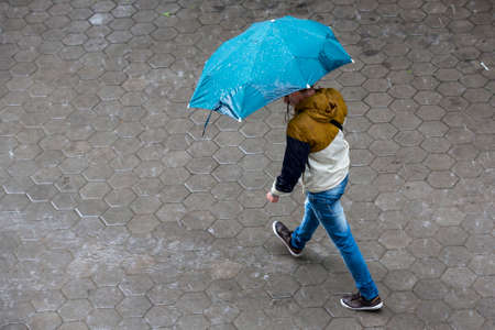 A man runs in the rainy weather with hir blue umbrella. Raining like cats and dogs. Bad weather. Climate change.の写真素材