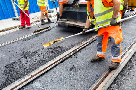 Team of workers put the hot asphalt on a street along tram car's railroad lines. Road construction workers with shovels in protective uniforms. Working in the hot day.の写真素材