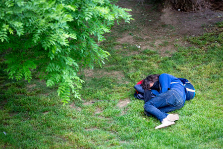 Homelsess person sleeps in a park under a tree with a face mask on.