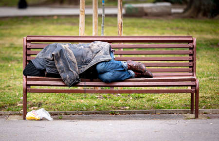 Homeless man sleeps on a bench covered with his jacket in a public park during the day.の写真素材