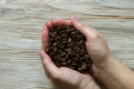 Roasted coffee beans in womans hands on wooden background, top view