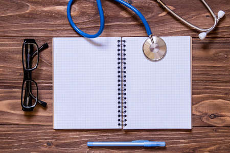 Opened notebook and other equipment such as pen, glasses, stethoscope on wooden office desk.の写真素材