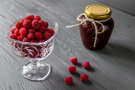 Fresh ripe raspberries in vintage crystal vase on old rustic wooden table. Jar of raspberry jam