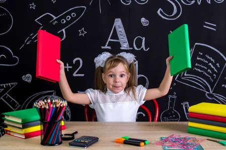 Happy schoolgirl sitting at the desk with books, school supplies, holding up two books in her both armsの写真素材