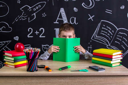 Smiling schoolboy sitting at the desk with a book before the face, hiding after the book, surrounded with school supplies. Chalkboard as a backgroundの写真素材