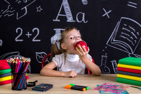 Hungry schoolgirl sitting at the desk with books, school supplies, holdingand biting the red appleの写真素材