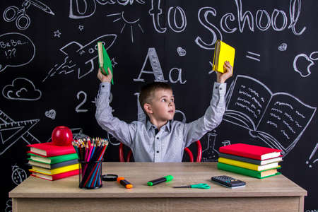 Schoolboy sitting at the desk with books, school supplies, holding up two books in his both arms, looking at themの写真素材