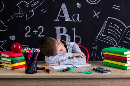 Schoolboy sleeping at the desk with a head leaned on the hands, surrounded with school supplies. Chalkboard as a backgroundの写真素材
