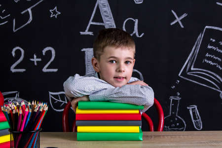 Excellent cute schoolboy sitting at the desk with a pile of books under the chin, surrounded with school supplies. Chalkboard as a backgroundの写真素材
