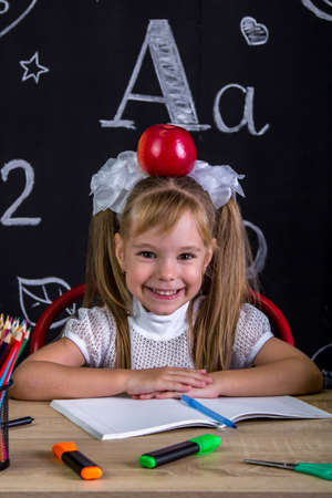 Cheerful and happy hellion schoolgirl sitting at the desk with books, school supplies, with a red apple on the top of her headの写真素材