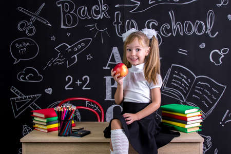 Cheerful schoolgirl sitting on the desk with books, school supplies, holding a red apple in the right handの写真素材