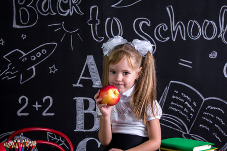 Greedy schoolgirl sitting on the desk with books, school supplies, yholding a red apple in the right...