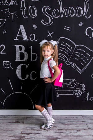 Coquette schoolgirl standing before the chalkboard as a background with a backpack on her backの写真素材