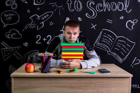 Unhappy youngster sitting at the desk with a pile of books under the chin, surrounded with school supplies. Chalkboard as a backgroundの写真素材