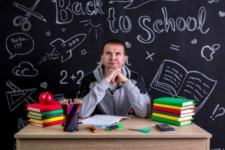 Disappointed and dissatisfied youngster sitting at the desk with a head leaned on the fists, surrounded with school supplies. Chalkboard as a backgroundの写真素材