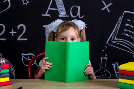 Schoolgirl sitting at the desk with a book before the face, hiding after the book, surrounded with...