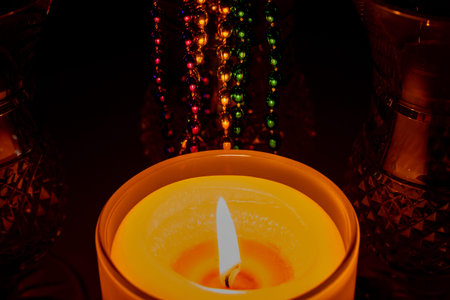 Macro abstract texture view of a glowing golden candle flame in a glass holder, illuminating nearby Mardi Gras beads in a darkened cornerの写真素材