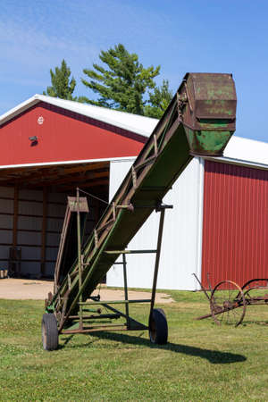 Close up abstract view of a weathered hay and grain elevator farm implement, with view of a large modern machine shed and blue sky backgroundの写真素材