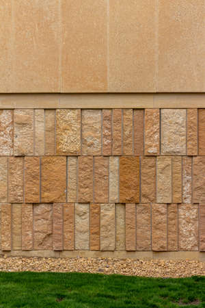 This image shows a rough textured limestone brick wall background with attractive vertical aligned natural stone blocks in shades of light brown, with view of foundation plants and grass.の写真素材