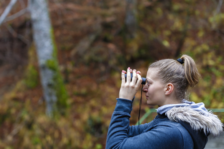 Young blonde woman tourist on a cliff looking through binoculars on the autumn landscape.の写真素材