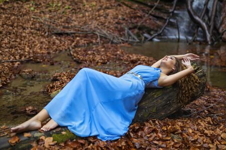 A beautiful woman in blue dress near a waterfall in autumn dayの写真素材