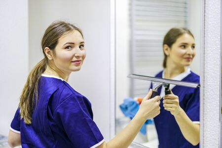 A girl in a cleaning service uniform cleans a mirror in the bathroom. The concept of cleanliness and disinfection.の写真素材
