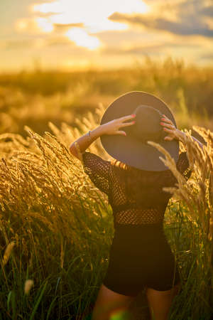 a beautiful brunette woman with hat poses in the grass at sunset.の写真素材