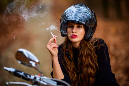 A beautiful long-haired woman smoking on a chopper motorcycle in autumn landscape on a forest roadの写真素材