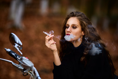 A beautiful long-haired woman smoking on a chopper motorcycle in autumn landscape on a forest roadの写真素材