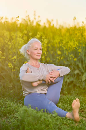 age woman relaxing on green grass during yoga exercise, morning routine, sunset light, outdoor. Mature female in sportswear doing stretching at nature open air. Age, healthy lifestyle.の写真素材