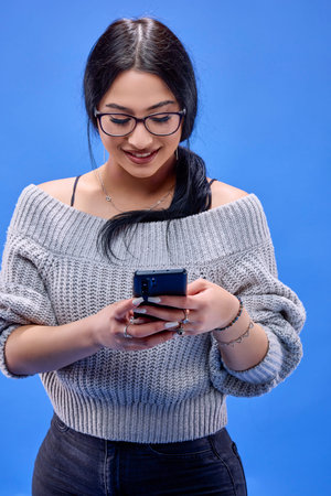beautiful young brunette with glasses posing in the studio on a blue background with a smartphone in...