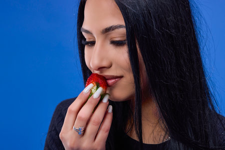 Portrait of a beautiful young brunette eating strawberries, isolated on a blue backgroundの写真素材