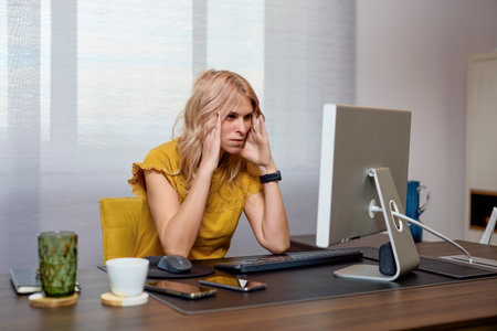 Tired businesswoman sitting at desk in office and looking at computer monitorの写真素材