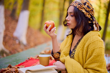 the beautiful brunette with a scarf on her head and a blanket is sitting at a table with a book, a cup and an apple in the autumn landscape.の写真素材