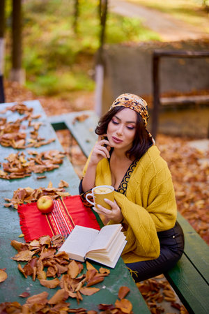 the beautiful brunette with a scarf on her head and a blanket is sitting at a table with a book, a cup and an apple in the autumn landscape.の写真素材