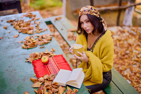the beautiful brunette with a scarf on her head and a blanket is sitting at a table with a book, a cup and an apple in the autumn landscape.の写真素材