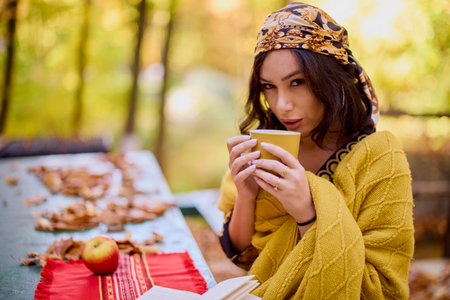 the beautiful brunette with a scarf on her head and a blanket is sitting at a table with a book, a cup and an apple in the autumn landscape.の写真素材