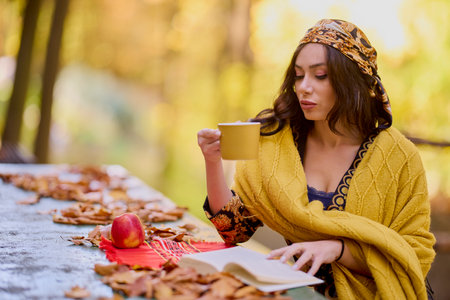the beautiful brunette with a scarf on her head and a blanket is sitting at a table with a book, a cup and an apple in the autumn landscape.の写真素材