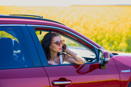 A beautiful woman with long hair stands next to a red SUV in a farm field.の写真素材