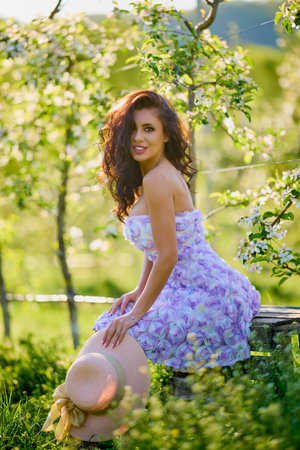 portrait of a beautiful woman in a dress sitting on a bench in a blooming apple orchard with a blurred backgroundの写真素材