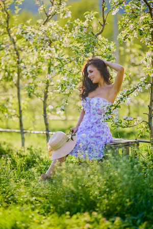 portrait of a beautiful woman in a dress sitting on a bench in a blooming apple orchard with a blurred backgroundの写真素材